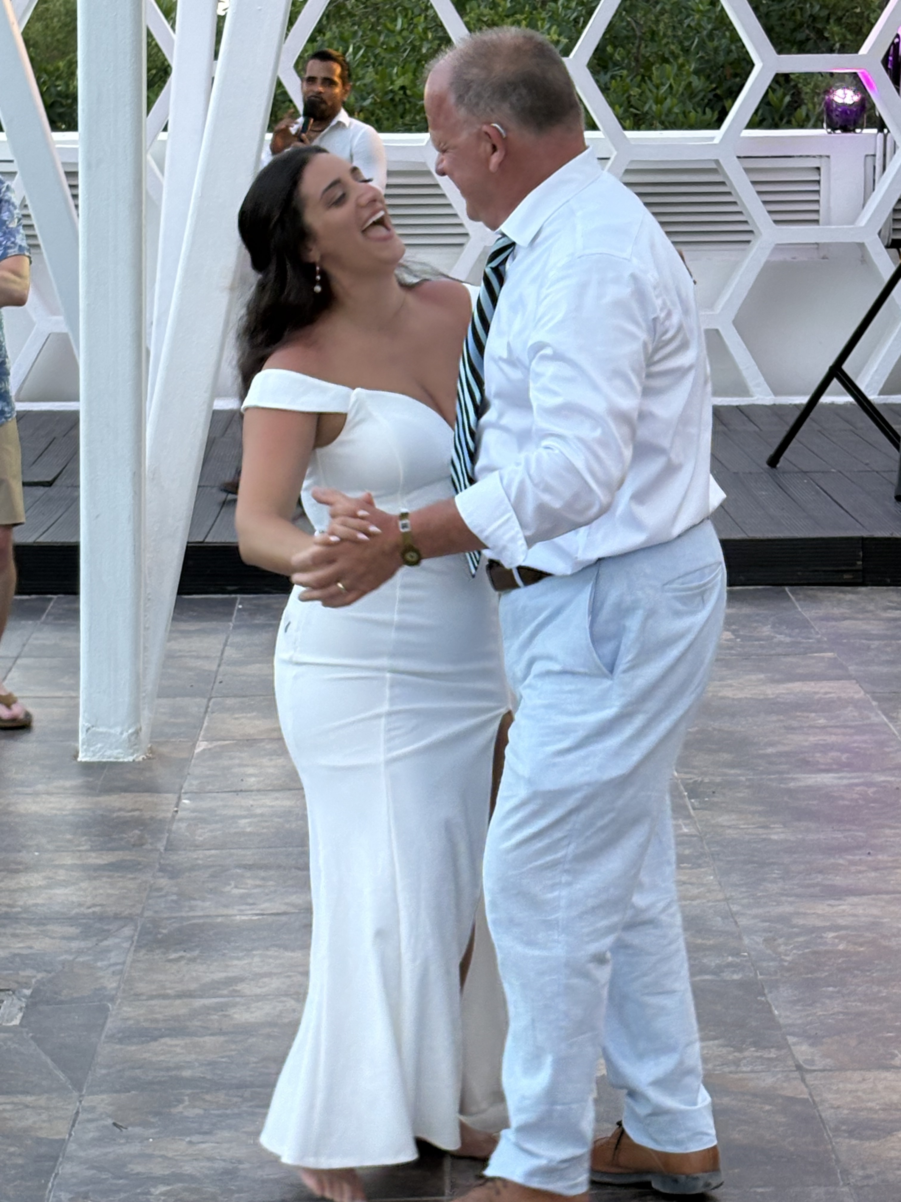 Aubrey and Noah share a joyful dance outdoors at their destination wedding, smiling together—she in a stunning white dress, he dressed smartly in a tie.