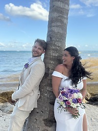 Aubrey and Noah: The bride and groom stand by a palm tree on the beach with a vibrant purple bouquet, capturing the essence of their beautiful destination wedding.