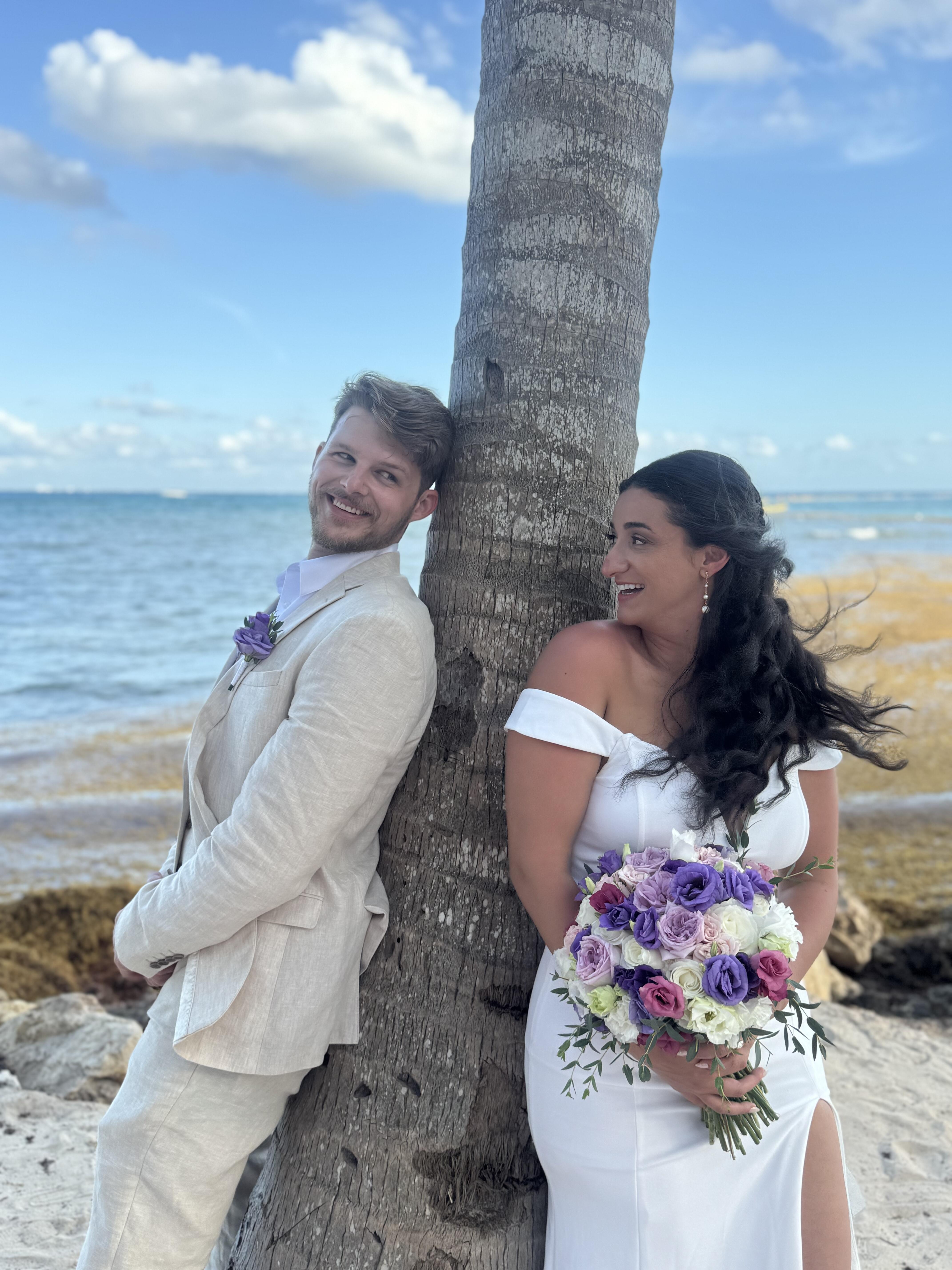 Aubrey and Noah: The bride and groom stand by a palm tree on the beach with a vibrant purple bouquet, capturing the essence of their beautiful destination wedding.
