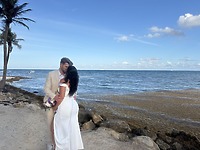 Sara and Tyler, dressed in their wedding attire, embrace on a rocky beach with the ocean providing a stunning backdrop to their destination wedding.