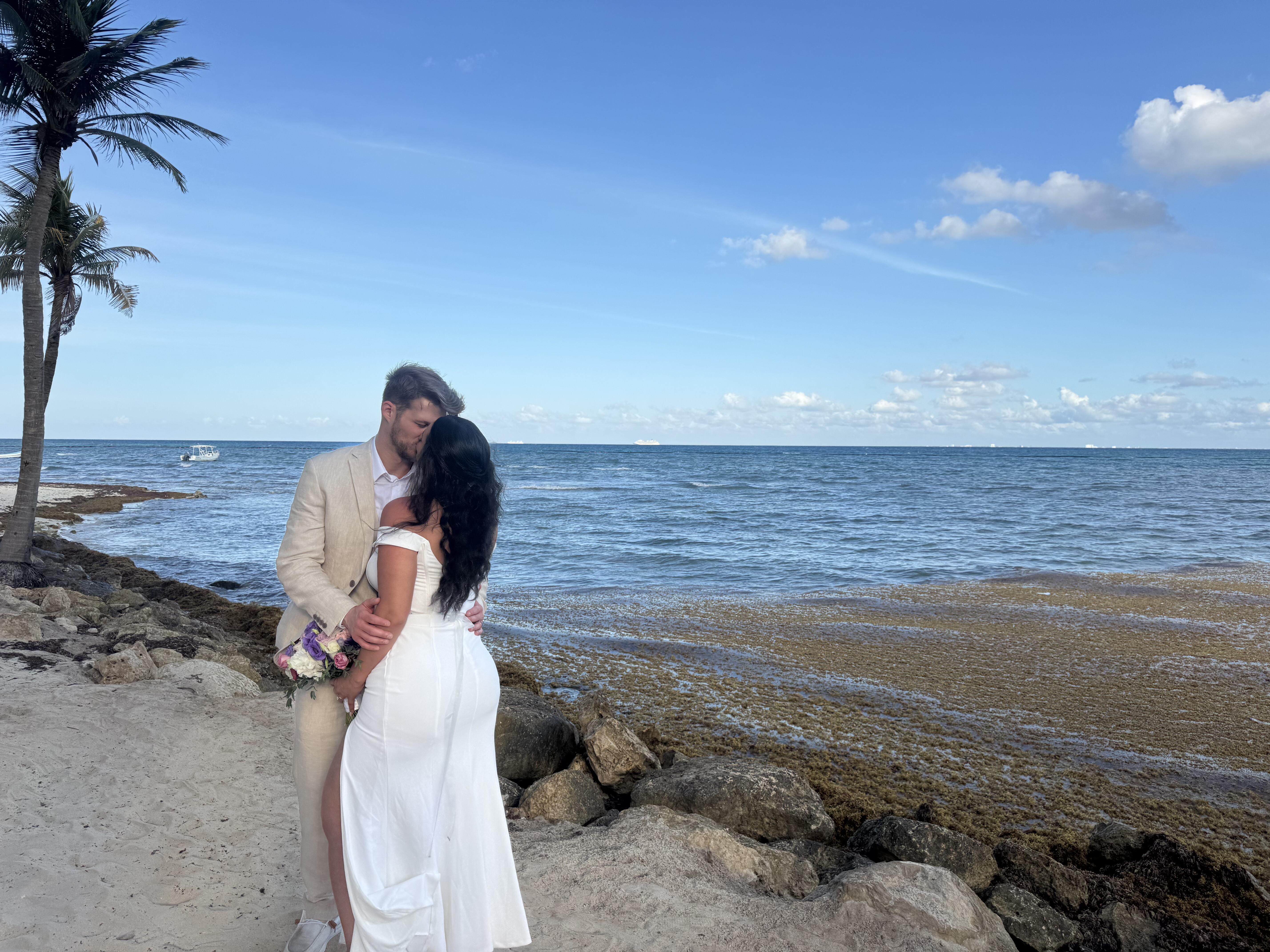 Sara and Tyler, dressed in their wedding attire, embrace on a rocky beach with the ocean providing a stunning backdrop to their destination wedding.