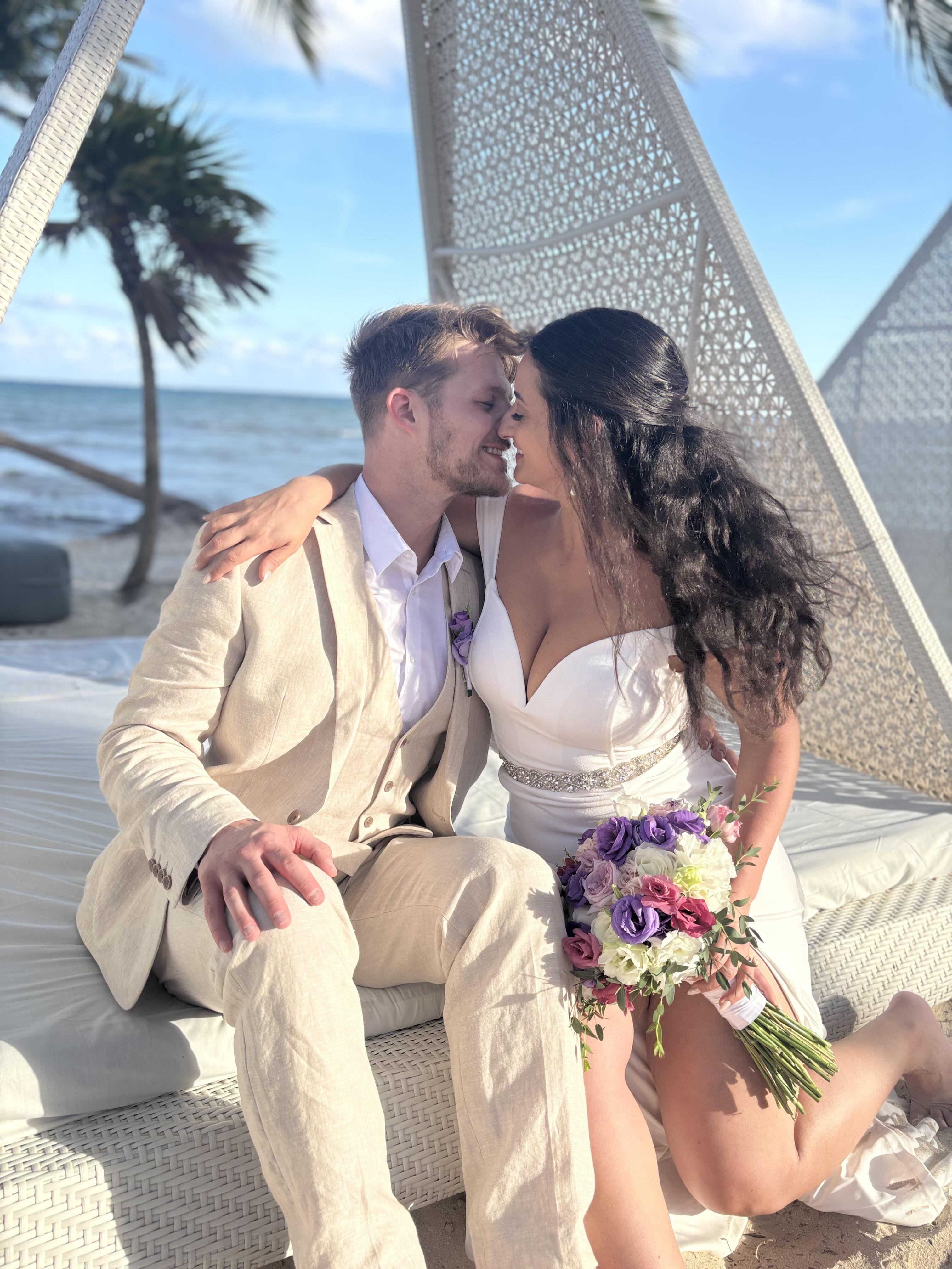 Sara and Tyler sit close together on the beach, smiling as they hold a bouquet, capturing a joyful moment from their destination wedding.