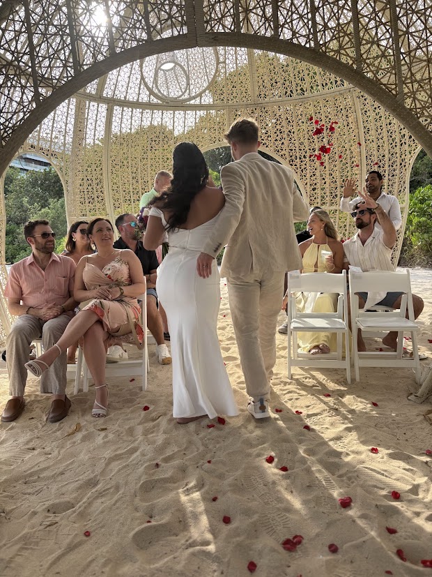 Aubrey and Noah walk down a sandy aisle at their destination wedding, while guests look on from beneath a beautifully decorated canopy.
