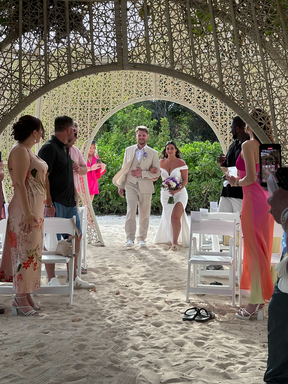 A bride and groom make their way down a sandy aisle at their destination wedding, passing beneath a woven arch while guests look on from either side.