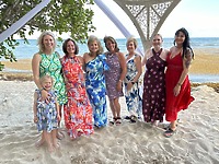 Eight women and a young girl, all dressed in Aubrey and Noah dresses, stand together on a sandy beach by the ocean, ready to celebrate a beautiful destination wedding.
