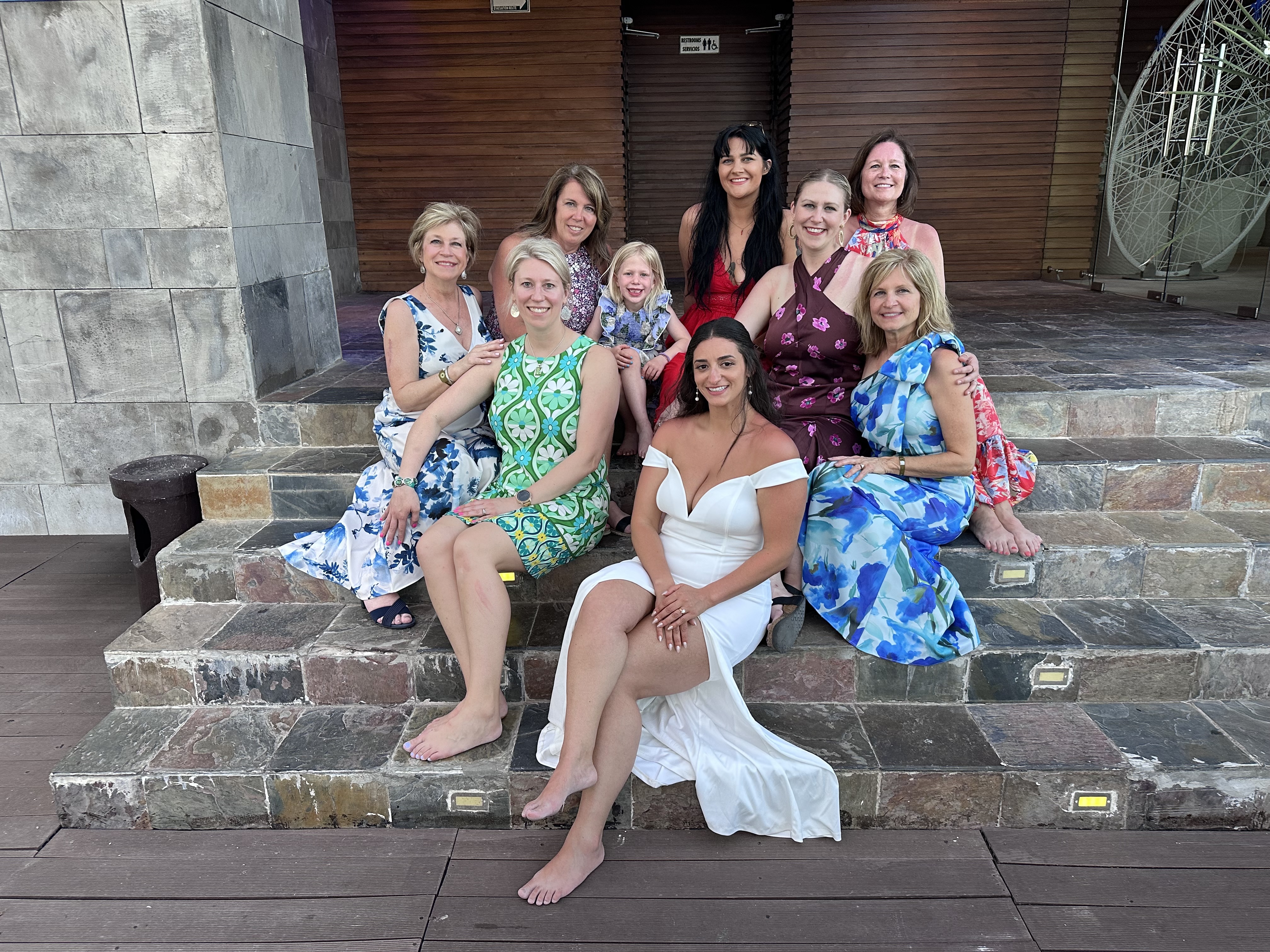 Nine women and a girl in colorful dresses sit on the steps, smiling together at the destination wedding—Aubrey and Noah’s celebration.