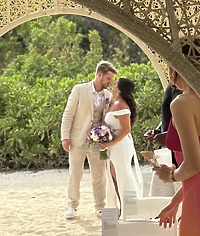 Aubrey and Noah stand together under a decorative arch on the sandy beach, celebrating their beautiful destination wedding.