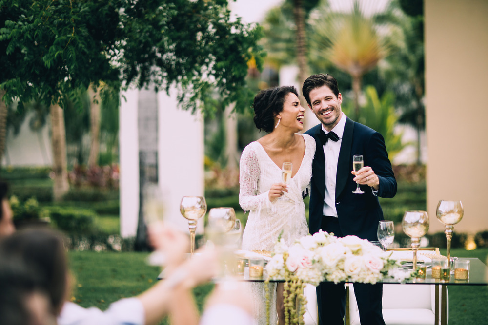 A bride and groom in elegant wedding attire smile and toast with champagne glasses at a beautiful outdoor destination wedding reception.