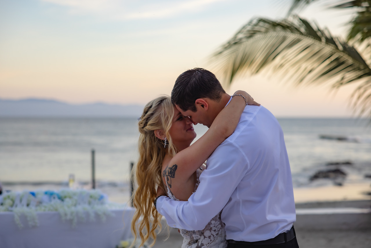 Camille and Jeff share a warm embrace on the beach at sunset, dressed in wedding attire, as they celebrate their destination wedding.