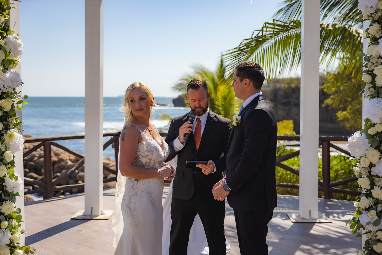 Camille and Jeff stand with the officiant during their outdoor destination wedding ceremony by the ocean.
