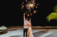 Camille and Jeff stand hand-in-hand on the beach at night during their destination wedding, pointing excitedly at the fireworks lighting up the sky.