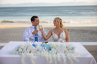 Camille and Jeff share a toast at their destination wedding, seated at a beachside table adorned with a “Mr & Mrs” sign.