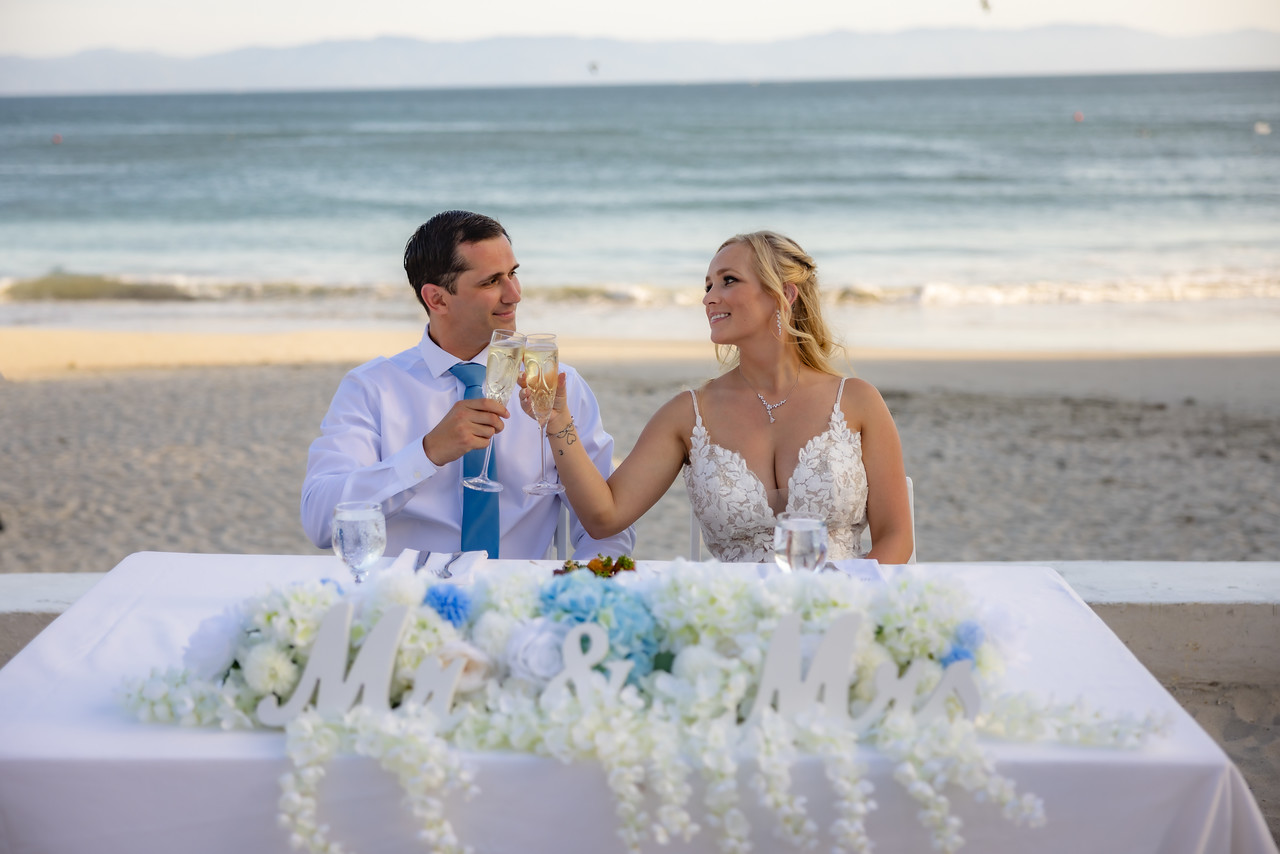 Camille and Jeff share a toast at their destination wedding, seated at a beachside table adorned with a “Mr & Mrs” sign.