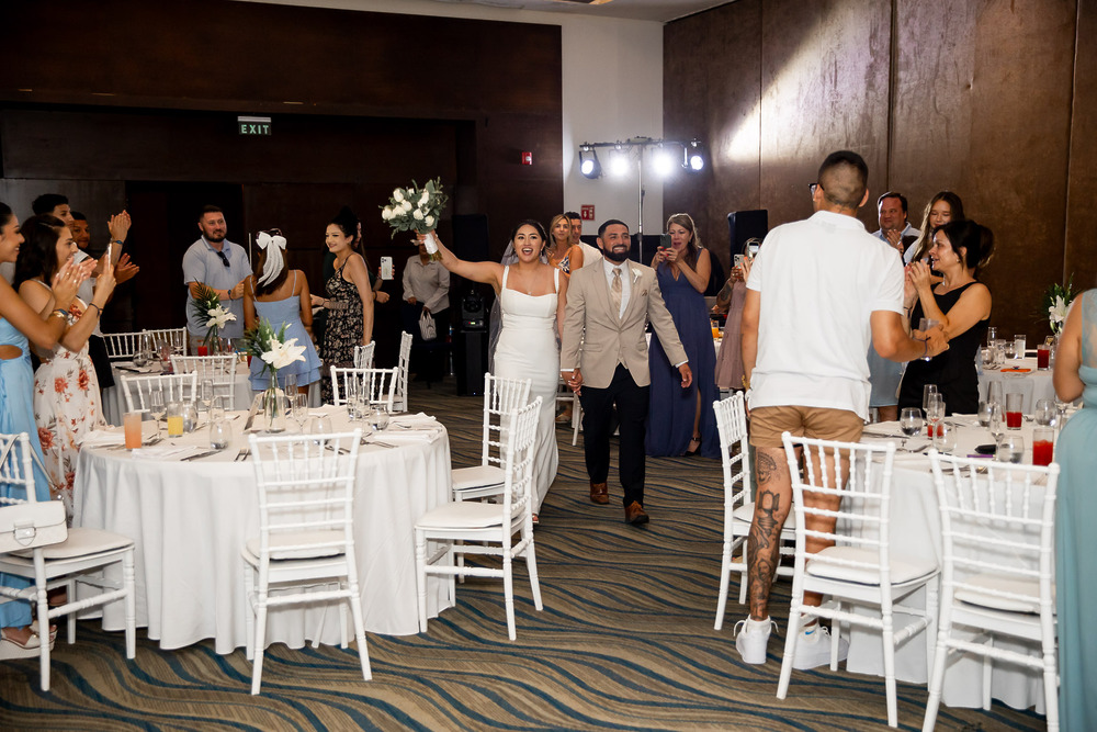 The newlywed bride and groom make a grand entrance into their destination wedding reception hall as guests cheer and applaud all around them.