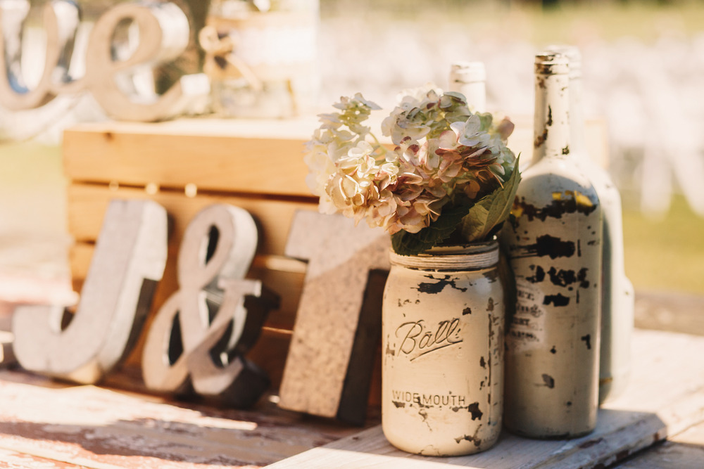 A rustic outdoor wedding table decorated with a mason jar filled with flowers, painted bottles, and wooden initials—perfect for a charming destination wedding.
