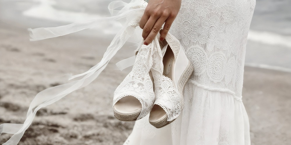 A person in a lace wedding dress stands on a sandy beach, holding white lace sandals, creating the perfect scene for a destination wedding.