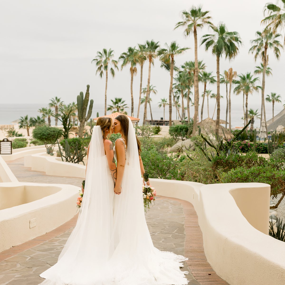 Two brides in white dresses share a kiss at their destination wedding, surrounded by palm trees and an ocean backdrop.