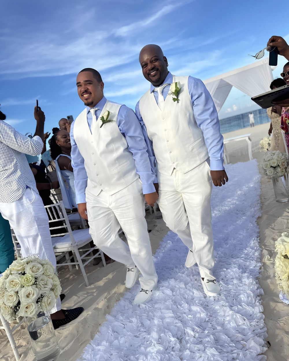 At their destination wedding, two grooms walk hand-in-hand down a flower-lined aisle on the beach.