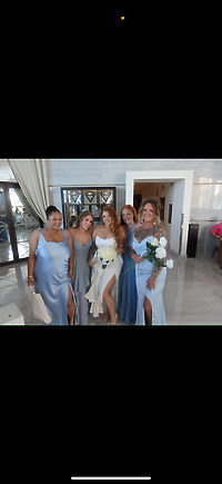 Five women pose indoors in elegant formal dresses at a destination wedding; one, dressed in a white Alexis and Colton gown, holds a bouquet.