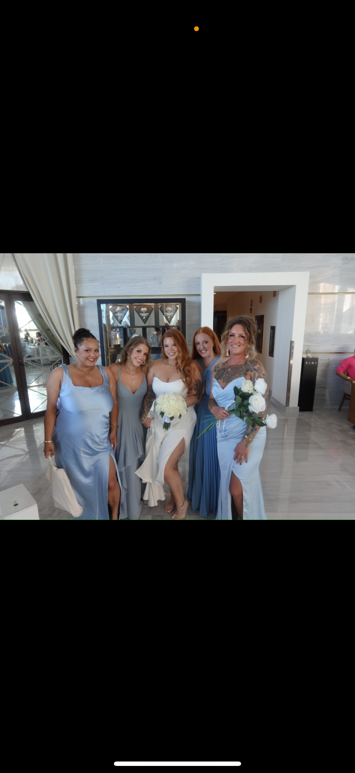 Five women pose indoors in elegant formal dresses at a destination wedding; one, dressed in a white Alexis and Colton gown, holds a bouquet.