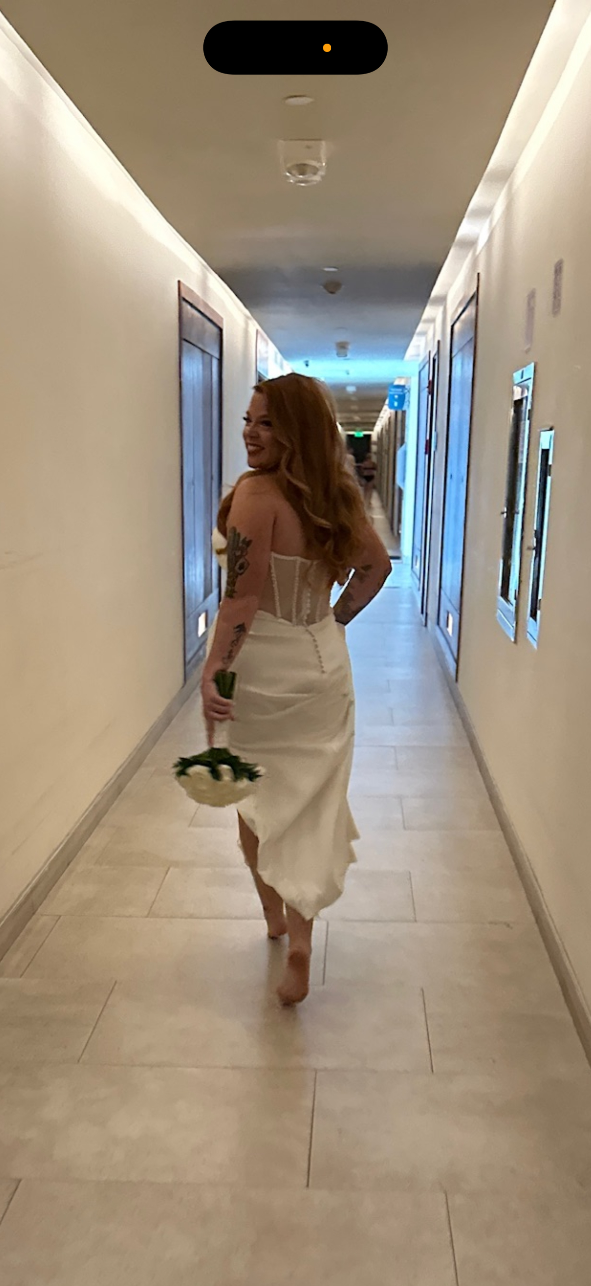 A woman, barefoot and smiling, walks down the hallway with flowers in hand, wearing the Alexis and Colton dress—radiating joy on her destination wedding day.