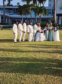 The Cynthia and Ruben destination wedding party poses outdoors on the grass, surrounded by palm trees and a picturesque building.