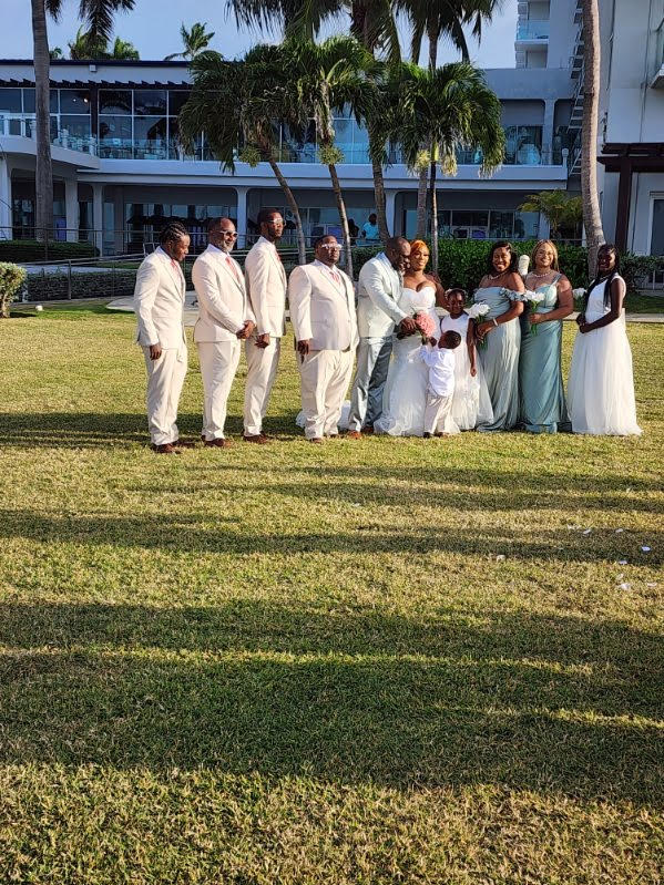 The Cynthia and Ruben destination wedding party poses outdoors on the grass, surrounded by palm trees and a picturesque building.