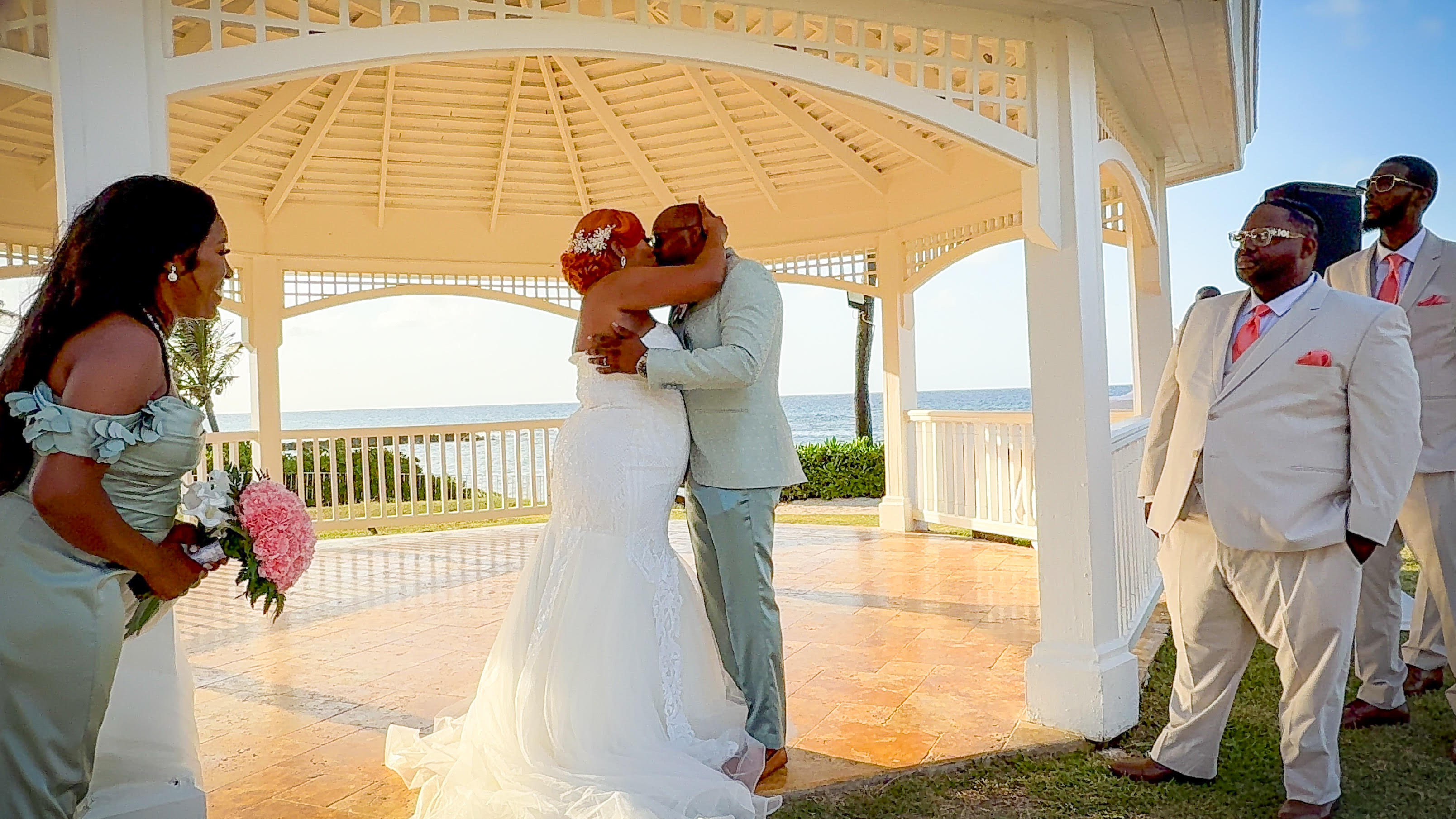 Cynthia and Ruben share an embrace beneath a gazebo as their wedding party stands nearby, capturing the joy of their beautiful destination wedding.