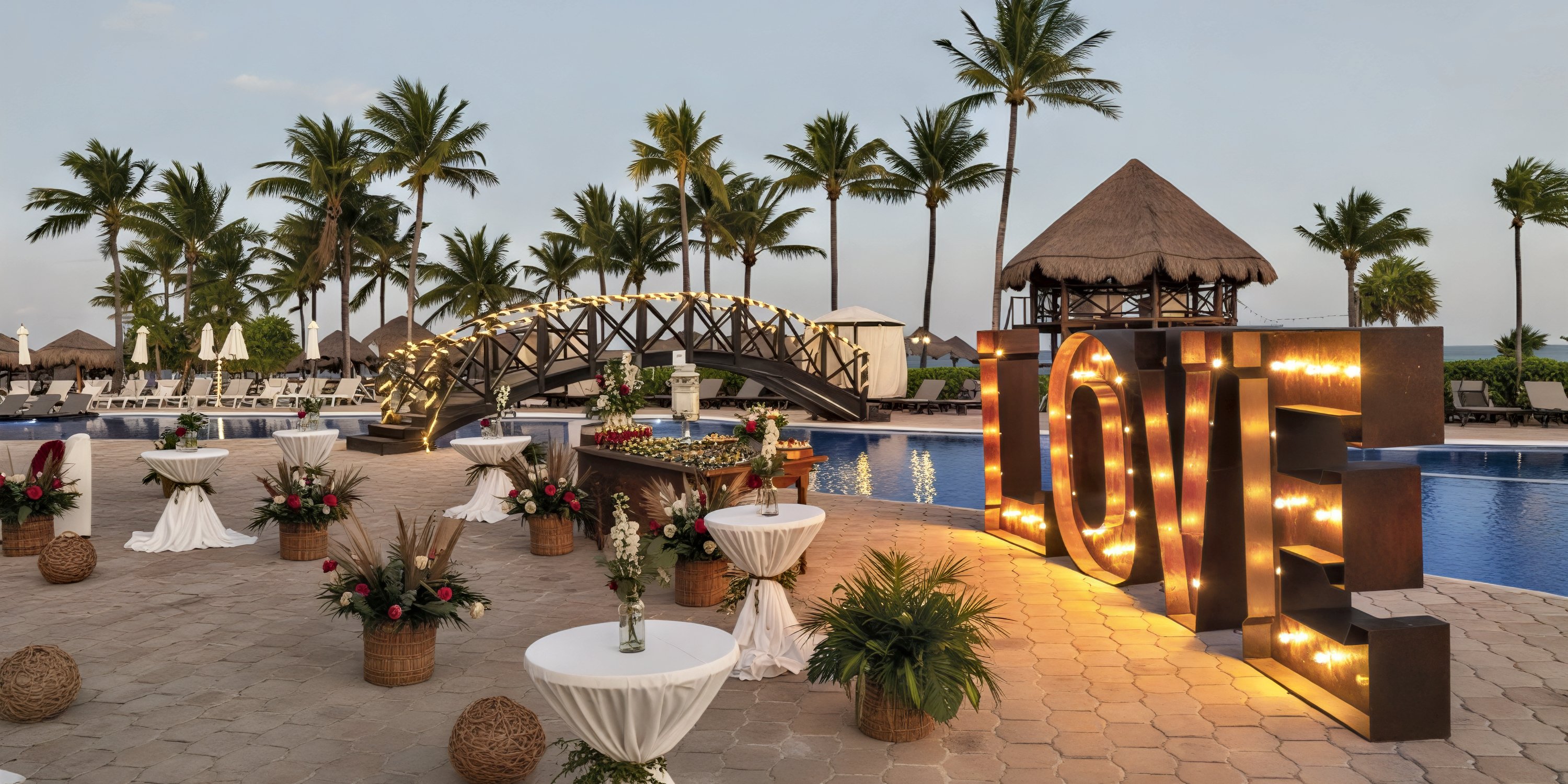 Ocean Maya Royale poolside wedding venue at sunset, featuring a "LOVE" marquee, cocktail tables, and swaying palm trees—the perfect setting for a destination wedding celebration.