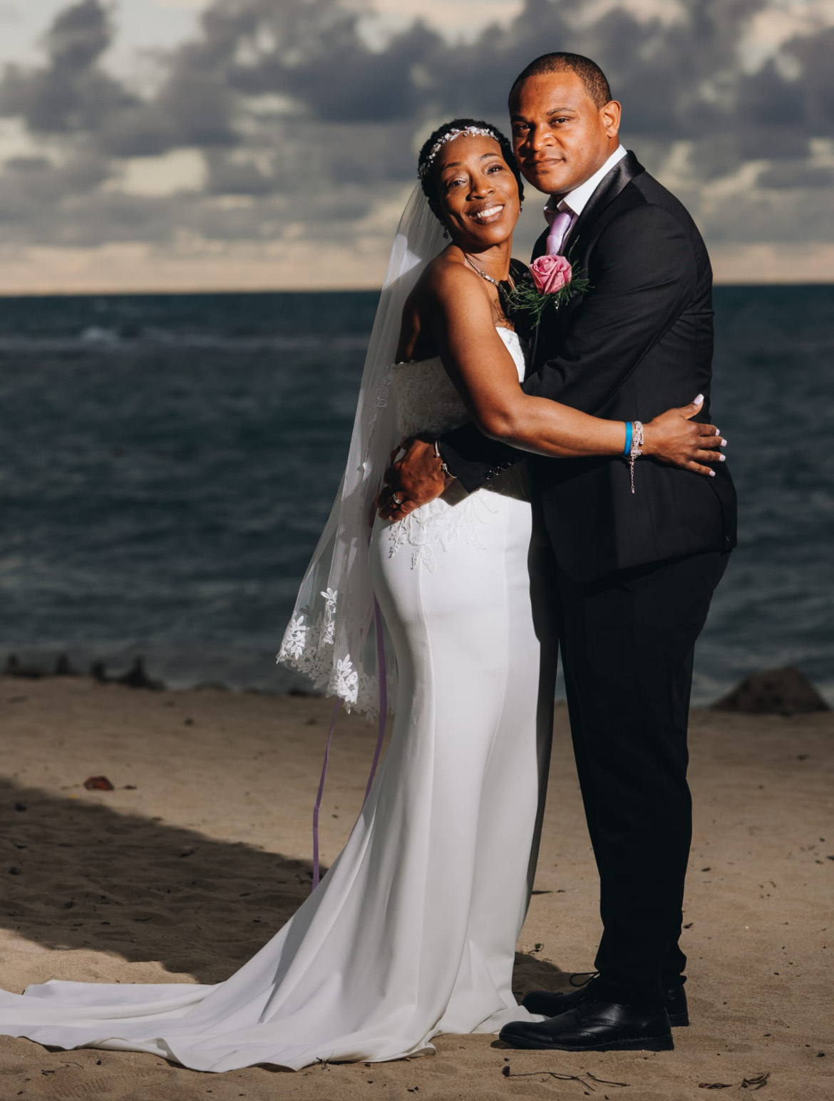 Natoya and Katrina embrace on a sandy beach with the ocean in the background, celebrating their destination wedding.