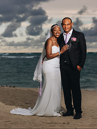 Natoya and Katrina stand side by side on a sandy beach, the ocean and a cloudy sky creating the perfect backdrop for their destination wedding.