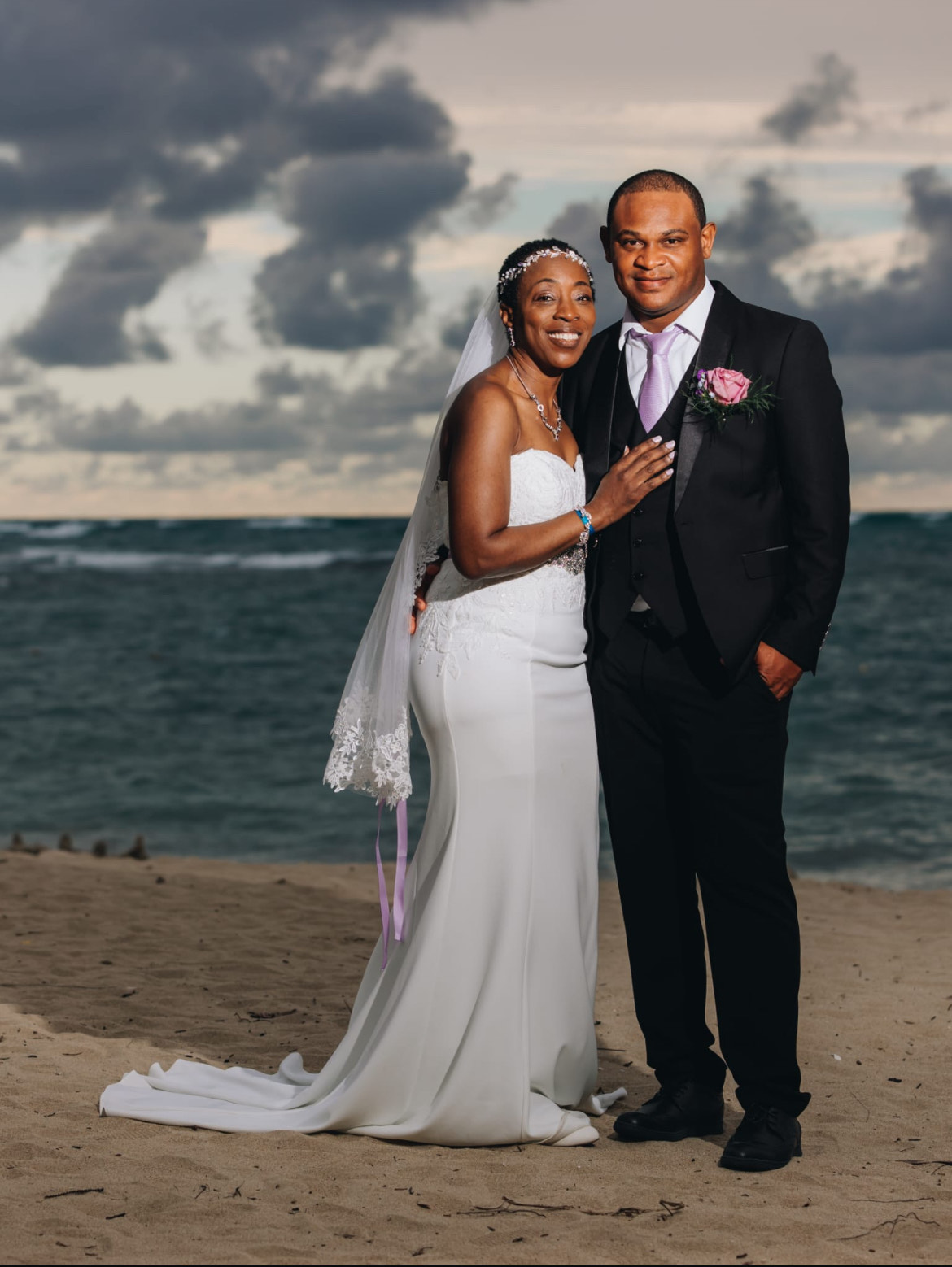 Natoya and Katrina stand side by side on a sandy beach, the ocean and a cloudy sky creating the perfect backdrop for their destination wedding.