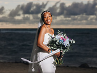Natoya and Katrina stand on a beach in white dresses, holding bouquets beneath a cloudy sky, celebrating their destination wedding.