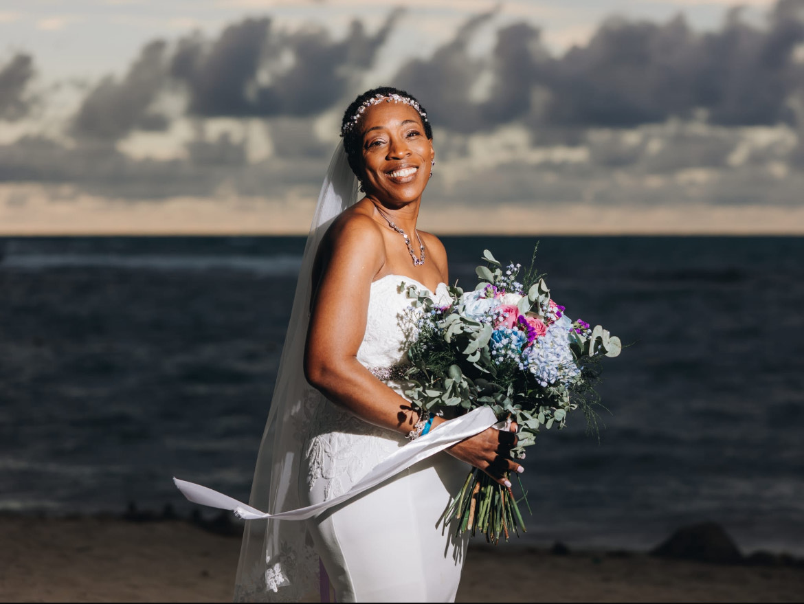Natoya and Katrina stand on a beach in white dresses, holding bouquets beneath a cloudy sky, celebrating their destination wedding.