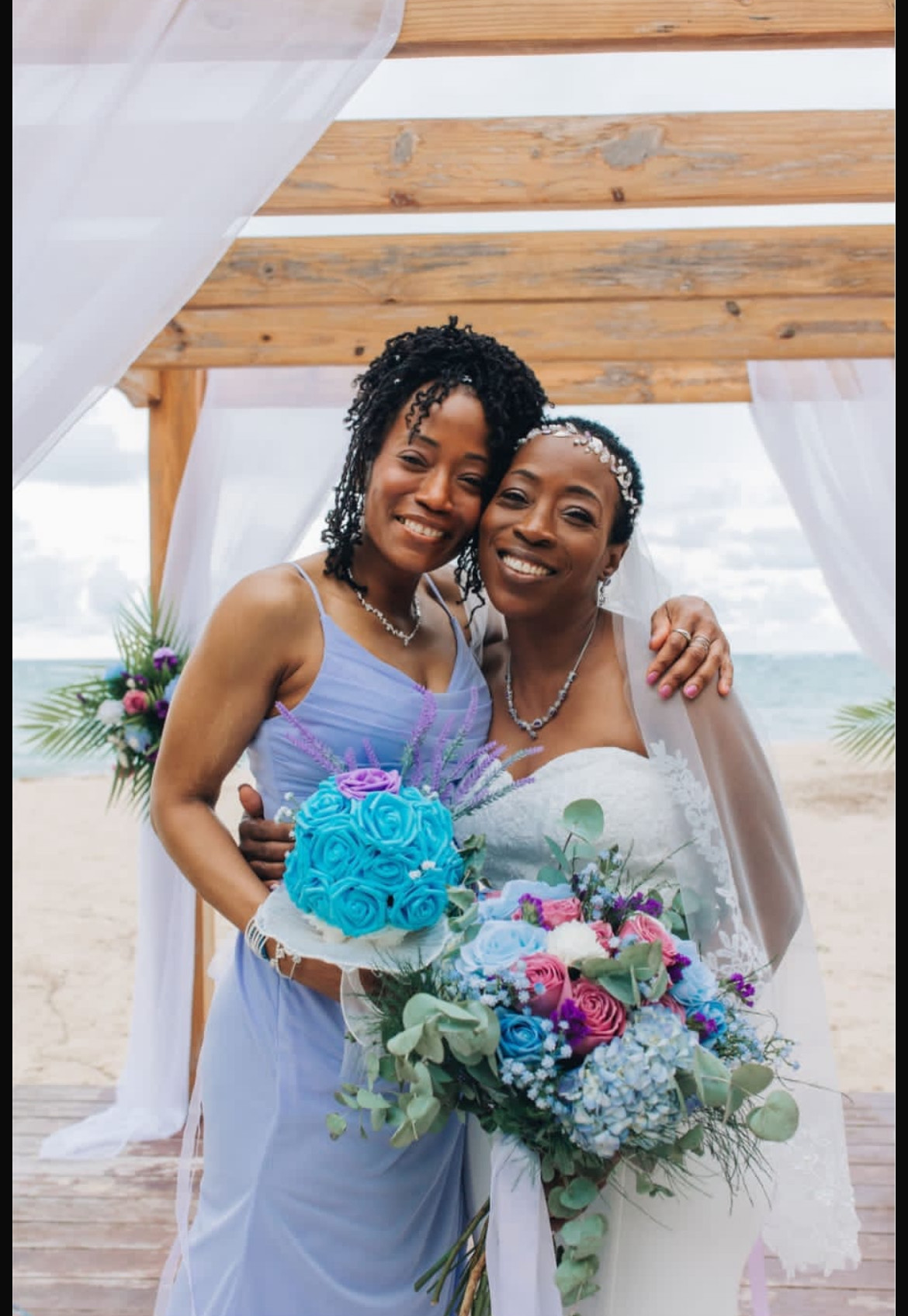 Natoya and Katrina at a destination wedding on the beach—one carrying a bouquet, the other holding a wedding cake.