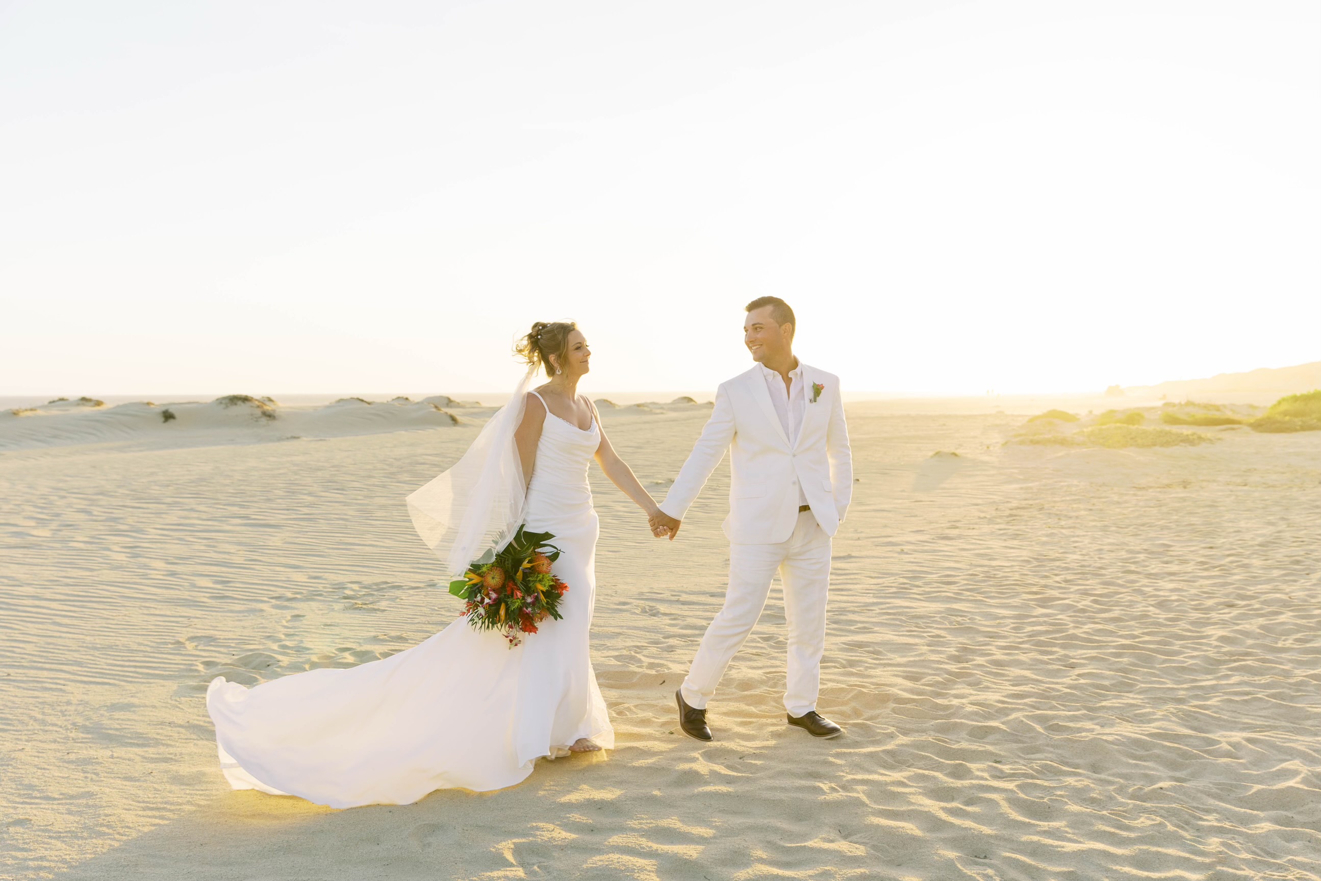 Alexis and Colton, dressed in white, walk hand-in-hand along the sandy beach at sunset, savoring a beautiful moment during their destination wedding.