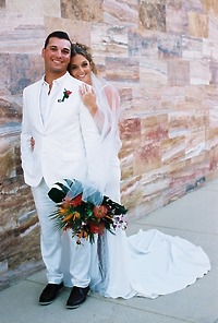 Alexis and Colton smile as they pose by a stone wall, holding a bouquet, capturing a joyful moment from their destination wedding.