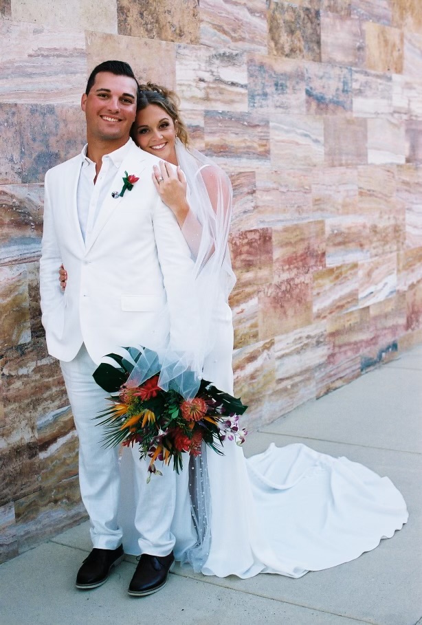 Alexis and Colton smile as they pose by a stone wall, holding a bouquet, capturing a joyful moment from their destination wedding.