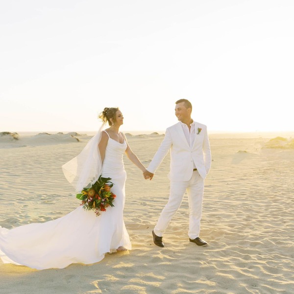 Alexis and Colton, dressed in elegant white wedding attire, walk hand-in-hand along the sandy beach at sunset, capturing the magic of their destination wedding.