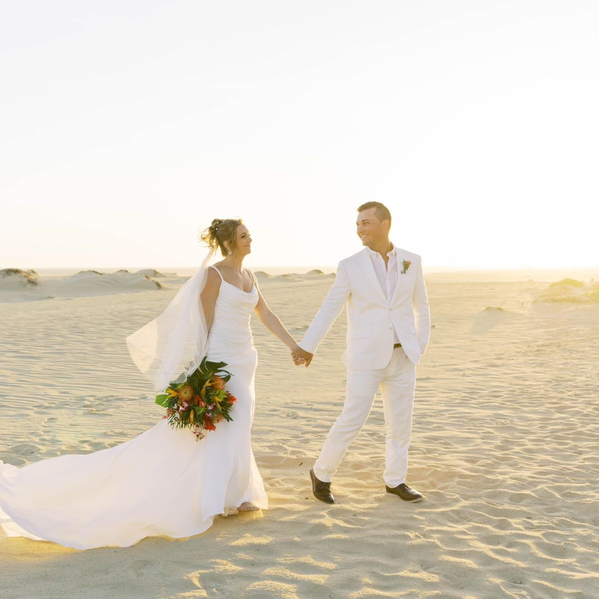 Alexis and Colton, dressed in elegant white wedding attire, walk hand-in-hand along the sandy beach at sunset, capturing the magic of their destination wedding.