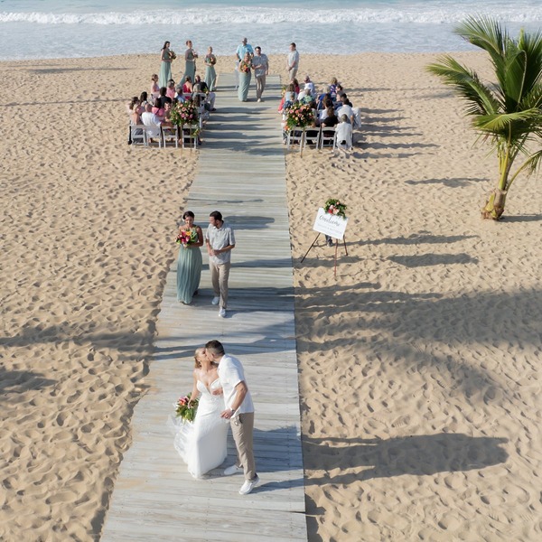 Tiana and Steven share a kiss on the boardwalk during their beautiful destination wedding, with guests seated behind them enjoying the romantic beachside ceremony.