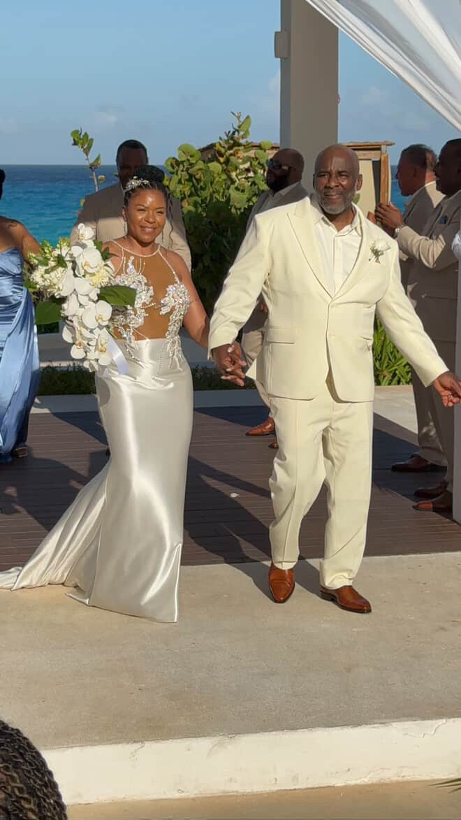 Khadijah and Oscar walk hand in hand, dressed in white, at their seaside destination wedding ceremony.