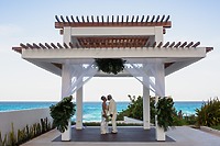 Khadijah and Oscar stand hand in hand under a white gazebo by the ocean, facing each other on their beautiful destination wedding day.