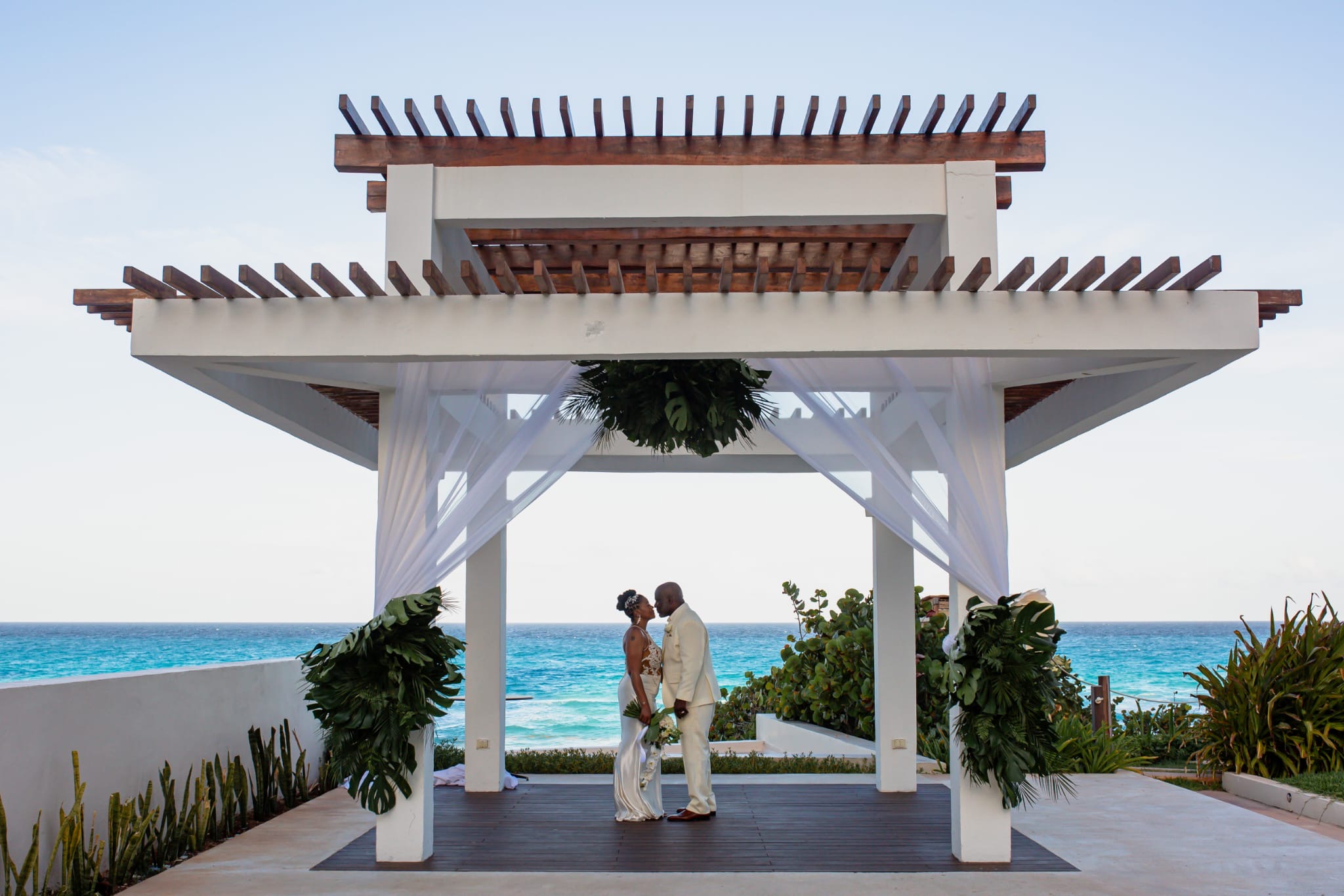Khadijah and Oscar stand hand in hand under a white gazebo by the ocean, facing each other on their beautiful destination wedding day.
