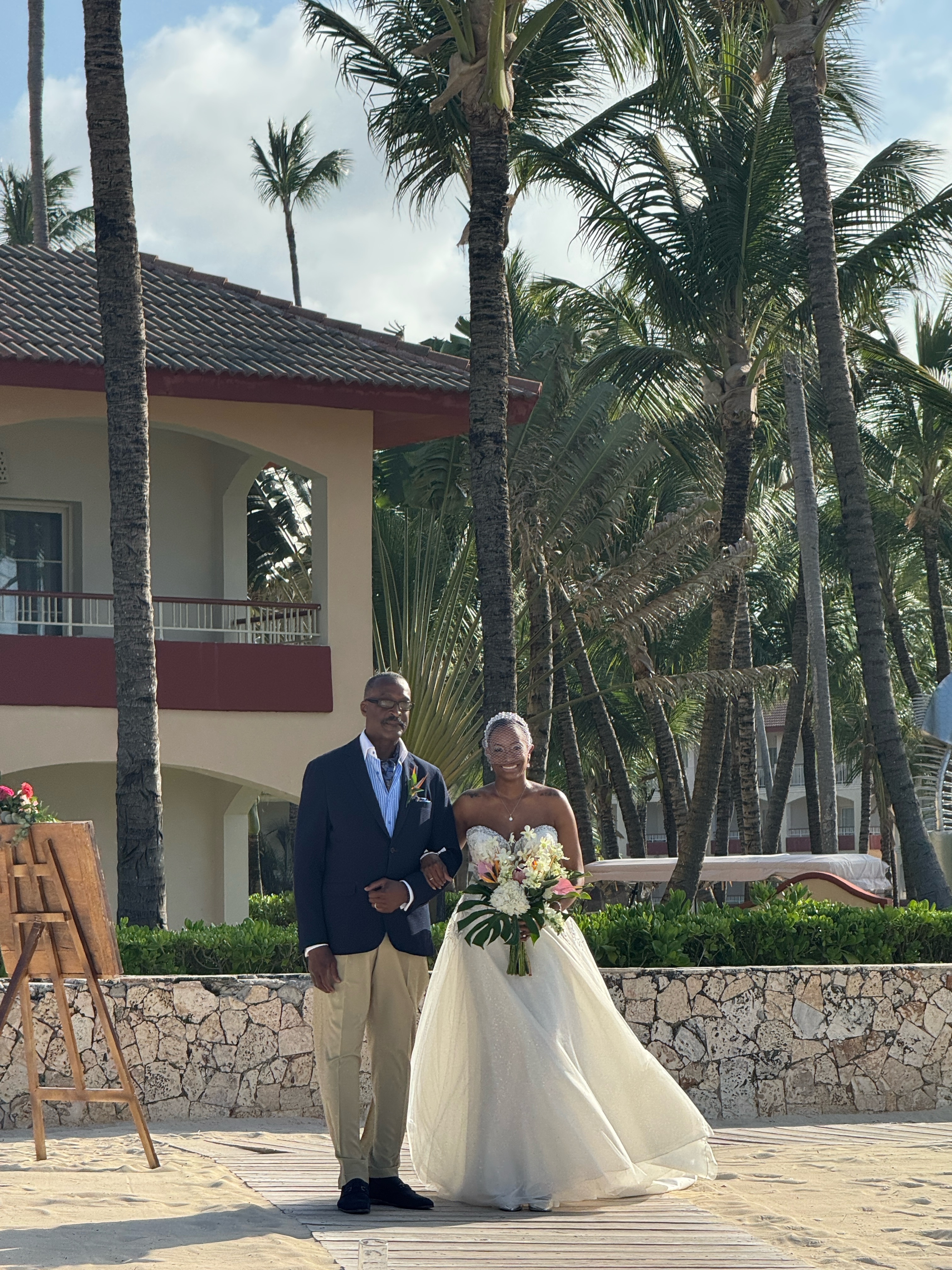 Jamie and Gary stroll outside near swaying palm trees and a luxurious villa, savoring the atmosphere of their destination wedding.
