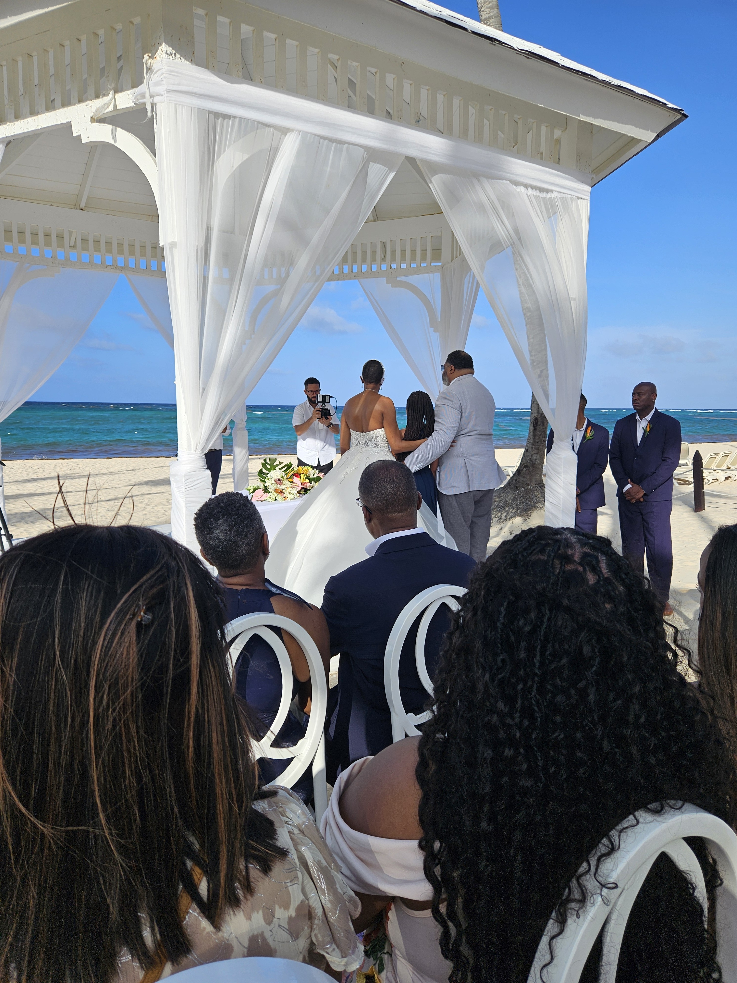 Jamie and Gary celebrate their destination wedding by exchanging vows at a scenic beach gazebo, with their guests seated nearby to witness the special moment.