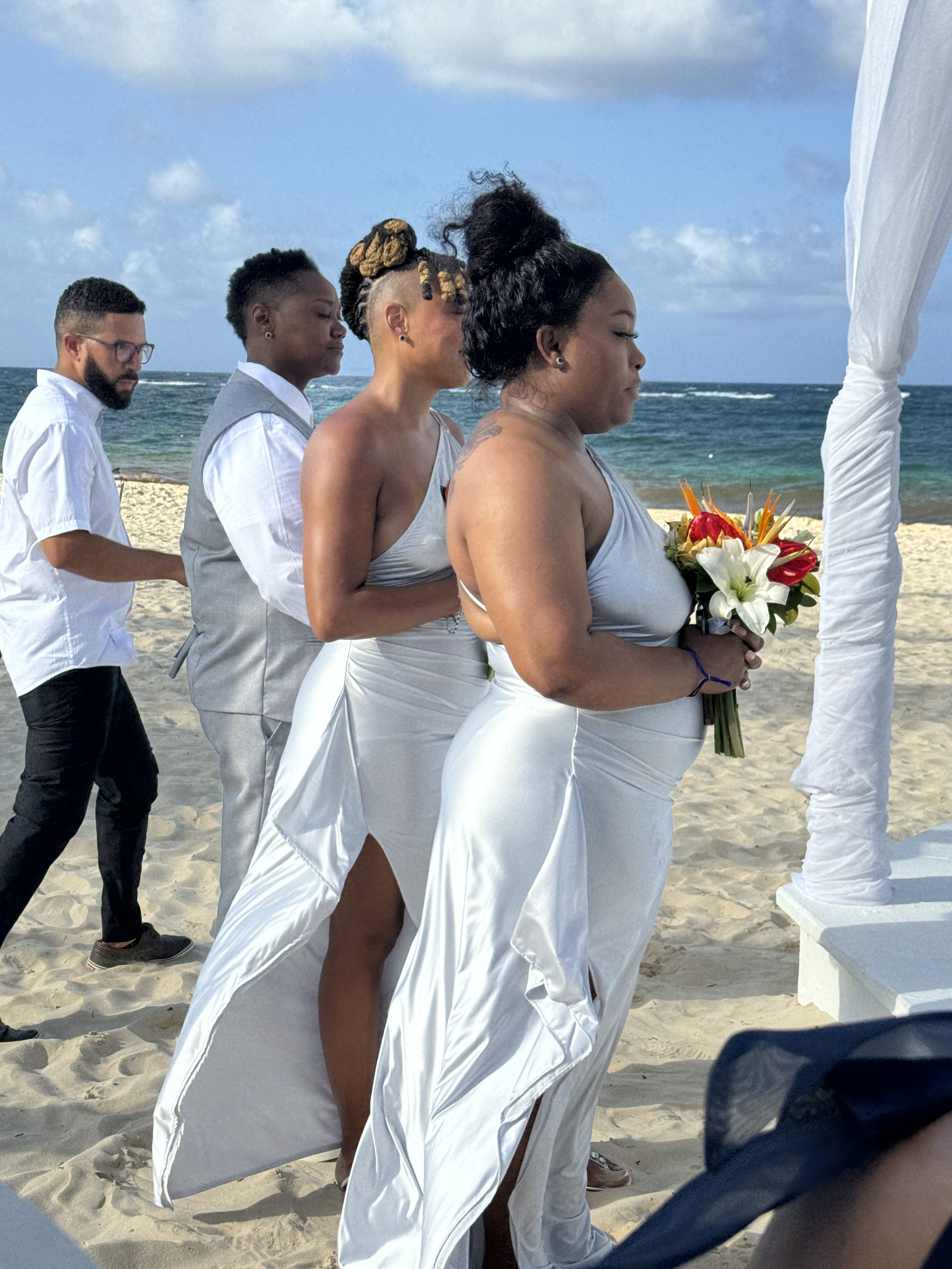 Three women in white dresses stand on the beach, celebrating Jamie and Gary's destination wedding.