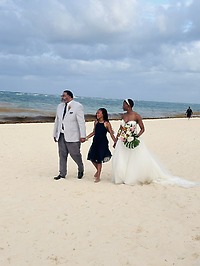 Jamie and Gary, along with a young girl, stroll along the sandy beach by the ocean at their destination wedding.