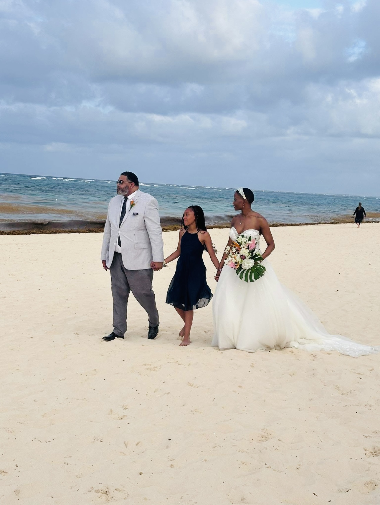 Jamie and Gary, along with a young girl, stroll along the sandy beach by the ocean at their destination wedding.