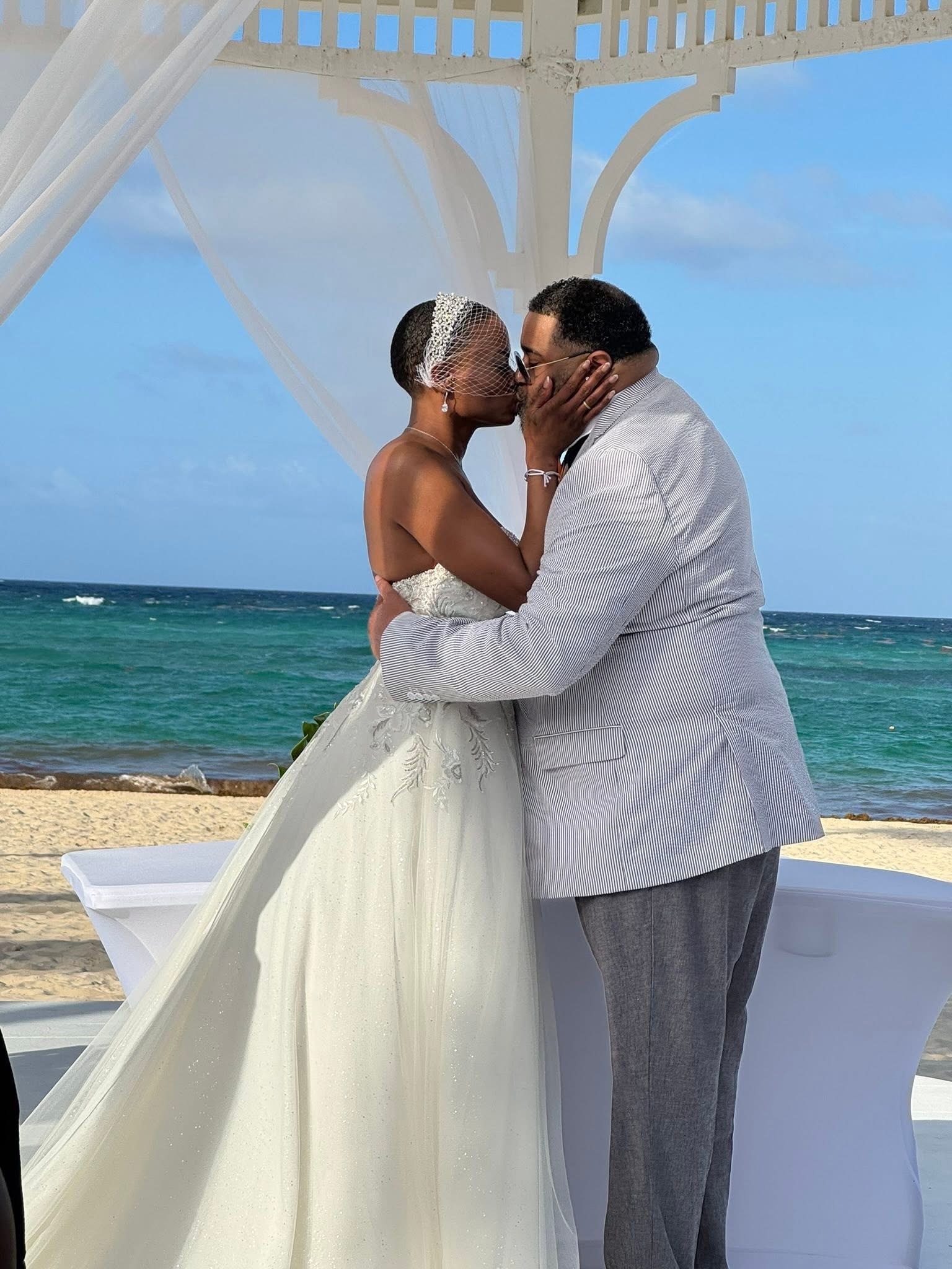 Jamie and Gary share a kiss beneath a white gazebo during their beautiful outdoor beach destination wedding ceremony.